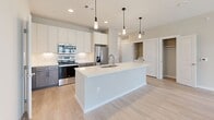 Located at 20130 Schiel Road in Cypress, Texas, this single-floor residence offers a thoughtfully designed open-concept layout with contemporary finishes throughout. The kitchen serves as the centerpiece of the living area, featuring a large center island with a white quartz countertop, stainless steel sink, and pendant lighting overhead. Cabinetry combines white upper cabinets with warm wood-toned lower cabinets accented by matte black hardware. Stainless steel appliances, including a refrigerator, range, and microwave, complete the modern kitchen. Light wood-look flooring extends throughout the main living areas, creating a bright and cohesive aesthetic. The living area opens to the outdoors through French doors flanked by windows, allowing abundant natural light and outdoor views. Bedrooms feature plush carpeting and windows with blinds that provide privacy while maintaining access to natural light. The primary bedroom includes a dedicated en-suite bathroom with a dual-sink vanity, white quartz countertops, dark wood cabinetry, large framed mirrors, and tile flooring. An in-unit laundry area is conveniently located just off the kitchen. The overall design palette of soft neutrals, clean lines, and modern hardware gives the entire space a fresh, move-in-ready appeal.