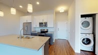 This single-floor residence showcases a thoughtfully designed open-concept layout that seamlessly connects the kitchen and living areas. The kitchen is a standout feature, outfitted with two-tone cabinetry — crisp white upper cabinets paired with slate gray lower cabinets — complemented by quartz countertops and a full suite of stainless steel appliances, including a refrigerator, electric range, and microwave. A central island with an undermount sink and pendant lighting above adds both functionality and style. Warm hardwood-style flooring runs throughout the main living areas, enhancing the contemporary aesthetic.

The living space is generously proportioned, bathed in natural light, and provides direct access to a private balcony through sliding glass doors. The bedrooms feature plush carpeting, ample closet space, and windows offering views of the surrounding building exterior. One bedroom includes a dedicated closet with built-in shelving.

A full bathroom is finished with a sliding-glass walk-in shower, hardwood-style flooring, and a dark-toned vanity. In-unit stacked washer and dryer hookups are conveniently accessible from the main living area. Recessed and flush-mount lighting throughout ensures a bright, clean atmosphere across all rooms.