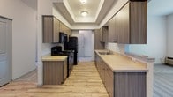 This single-floor residence offers a well-organized layout across several distinct living spaces. The galley-style kitchen is a functional highlight, featuring warm wood-toned cabinetry on both walls, light-colored countertops, and black appliances including a gas range and refrigerator. A tray ceiling with recessed lighting adds architectural interest overhead, while light hardwood-style flooring runs the length of the kitchen, leading toward additional living areas.

The bedrooms are carpeted in a neutral grey tone and share a consistent design language of clean white walls and natural light provided by window blinds. Each bedroom includes a sliding-door closet with a hanging rod, offering practical storage. Views from the bedroom windows look out toward surrounding greenery and neighboring structures.

The bathroom is finished with large-format tile flooring in warm earth tones, providing a durable and visually grounding surface. A single-sink vanity features cabinetry that mirrors the kitchen's wood-tone finish, complemented by a wide mirror and light fixture above. A full-size tub and shower combination with a curtain rod completes the bathroom.

The overall ambiance of the space is clean, modern, and neutral, making it a functional and move-in ready environment.