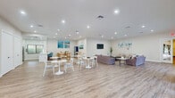 This single-floor community clubhouse is designed with an open, airy layout that promotes social gathering and relaxation. The main gathering area is expansive, featuring light wood-look flooring, recessed lighting, and crisp white walls that create a clean, modern atmosphere. Multiple round white bistro tables with matching chairs are arranged throughout the space, offering flexible seating for casual meetings or social events. A comfortable lounge area is furnished with plush sofas and accent chairs, centered around a wall-mounted flat-screen television, and accented with coastal landscape artwork and potted greenery that lend a serene, resort-like feel. A dedicated dining nook features natural wood furniture, a sideboard, a navy display cabinet, and additional coastal-themed artwork, adding warmth and character to the space. The fully equipped kitchen includes white shaker cabinetry, quartz countertops, stainless steel appliances, a decorative tile backsplash, and a large island, all illuminated by recessed lighting and a window overlooking the outdoors. The reception entry area offers a front desk with a package room, wood flooring, and direct access to the exterior through both a single door and elegant French doors that invite natural light deep into the space.