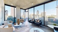 This single-floor space showcases a sophisticated industrial-modern design, defined by exposed concrete ceilings and expansive floor-to-ceiling glass walls that frame striking panoramic views of a dense urban skyline. The open-plan living area centers around large-format tile flooring, with seating arrangements that include sleek black leather sofas, contemporary lounge chairs, and round white coffee tables atop layered area rugs—creating distinct zones for conversation and relaxation.

Natural light floods the interior through the wraparound glazing system, reinforcing a seamless visual connection between indoors and out. One interior wall features a mounted flat-screen television flanked by framed artwork, balanced against crisp white walls that contrast the raw concrete overhead.

The space extends to multiple outdoor terrace areas. One rooftop terrace offers a fire pit centerpiece surrounded by cushioned lounge seating and unobstructed 360-degree skyline views behind glass railings. A second terrace features an intimate courtyard-style layout with raised planters of manicured greenery, additional lounge seating, and a fire feature, providing a more sheltered al fresco setting. Together, the indoor and outdoor areas create a cohesive, elevated entertaining environment with a distinctly urban character.