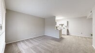 This thoughtfully laid-out one-bedroom, one-bathroom residence makes efficient use of its space across a single floor. The open-concept living and kitchen area serves as the heart of the home, with plush carpeting in the living room flowing seamlessly toward the kitchen. The kitchen features crisp white shaker-style cabinetry, marble-look countertops, a full suite of black appliances including a refrigerator, range, microwave, and dishwasher, and light wood-look flooring that adds warmth and contrast. A pass-through counter connects the kitchen to the living area, enhancing the open feel of the shared space. The bedroom offers generous proportions, carpeted flooring, natural light from a window with blinds, a ceiling fan with light fixture, and direct access to a dedicated closet with wire shelving. A second closet provides additional storage. The full bathroom is finished with a white shaker vanity topped with a marble-look surface, a large mirror, wood-look flooring, and a tiled shower-tub combination. Neutral warm-gray walls throughout the residence create a calm, cohesive aesthetic, while an entry area offers a practical transition into the living spaces.