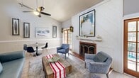 Located at 7502 Hunt Club Rd in Columbia, South Carolina, this single-floor space offers an efficient yet thoughtfully designed open-concept layout combining the kitchen and living area into one cohesive space. The living room is anchored by a wood-burning fireplace framed by a striking white shiplap accent wall with diagonal paneling, adding a bold architectural focal point. Vaulted ceilings enhance the sense of openness, while a ceiling fan ensures year-round comfort. Light-toned hardwood-style flooring runs throughout, complementing the soft gray walls accented by classic white wainscoting. French doors flood the space with natural light and offer a connection to the outdoors. A compact dining area with a round table and modern chairs tucks neatly alongside the living space. The kitchen features a peninsula with a white quartz-style countertop and bar seating, gray shaker cabinetry, a geometric tile backsplash, and black appliances. Shiplap detailing on the kitchen peninsula ties together the cohesive farmhouse-modern aesthetic present throughout. The overall ambiance is warm, clean, and inviting, blending contemporary design elements with cozy, character-rich details.