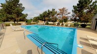 This community amenity space includes two distinct areas: an outdoor swimming pool and an attached support building housing a shared laundry facility and individual storage units. The outdoor pool is generously sized, filled with clear turquoise water, and features lane dividers for organized use. Multiple entry points with metal handrails provide safe access, and the surrounding concrete deck is lined with lightweight lounge chairs, offering a comfortable space to relax. Mature trees and landscaped greenery frame the area, with views of the broader community visible in the background.

The support building is accessed through a dark-painted entry door with a glass panel and contains a well-organized laundry room equipped with multiple top-loading washers and dryers. The room features light-colored walls, vinyl flooring with a blue accent mat, overhead fluorescent lighting, and a folding table for convenience. Posted informational notices are visible on the walls. Adjacent to the laundry area, a narrow hallway with overhead recessed lighting leads to a row of individual storage lockers on both sides, providing additional storage options for residents.