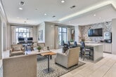 Located at 2370 Main St NW in Duluth, Georgia, this sophisticated single-floor space serves as a polished community or leasing center with a refined, welcoming atmosphere. The interior is finished with large-format light stone tile flooring throughout, creating a clean and cohesive foundation that ties each area together seamlessly.

The main lounge area is anchored by a substantial marble-topped counter with navy blue upholstered bar seating, complemented by neutral-toned armchairs arranged around a stone coffee table atop a patterned area rug. Expansive arched glass windows and glass-paneled sliding doors flood the space with natural light and offer views of the surrounding exterior, lending an open and airy quality to the interior.

A dedicated workstation area features a large farmhouse-style wooden table set beneath a candle-style chandelier, surrounded by dark leather chairs and positioned beside tall arched windows that bring in abundant daylight and outdoor views of the community.

The kitchenette and media area is highlighted by a bold herringbone-patterned tile accent wall framing a wall-mounted flat-screen television, white cabinetry, and quartz countertops with a built-in sink and coffee station. Curated artwork and potted greenery add warmth and character throughout the space, while recessed lighting provides consistent, well-distributed illumination across all areas.