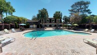 Located at 711 Underwood Avenue in Pensacola, Florida, this community amenity space offers a welcoming resort-style atmosphere designed for relaxation and outdoor enjoyment. The centerpiece of the space is a generously sized, kidney-shaped swimming pool with crystal-clear turquoise water, framed by an expansive paver brick deck that provides ample room for lounging and socializing. Colorful patio umbrellas and outdoor seating arrangements are positioned throughout the deck, offering shaded areas perfect for gathering under the Florida sun. Tall, mature palm trees and neatly manicured shrubbery frame the space, lending a lush, tropical character to the setting. The backdrop features a charming two-story clubhouse building with a classic architectural style, highlighted by a dark mansard-style roof, white-trimmed windows, and a raised entrance flanked by well-maintained hedges and greenery. Additional seating areas with vibrant umbrellas extend along both sides of the pool, enhancing the social and recreational appeal. The overall ambiance is bright, open, and inviting, reflecting the quintessential outdoor lifestyle of the Florida Gulf Coast region.