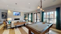 This well-appointed resident room offers a thoughtfully designed, multi-functional space ideal for socializing, recreation, and focused work. The open-concept lounge area serves as the centerpiece, anchored by a striking floor-to-ceiling stacked stone accent wall housing a sleek linear fireplace and a large wall-mounted television. Comfortable seating arrangements, layered with patterned rugs, warm accent lighting, and vibrant teal drapery panels, create an inviting and contemporary atmosphere. Natural light pours in through full-length glass French doors that open to an outdoor terrace, enhancing the airy feel of the space. Rich hardwood-style flooring in a light, wide-plank finish runs throughout, adding warmth and visual continuity. A full-size billiards table with pendant lighting above occupies one side of the lounge, offering a leisure focal point for residents. The kitchenette is fully equipped with dark granite countertops, taupe shaker-style cabinetry, a stainless steel refrigerator, dishwasher, microwave, sink, and a coffee station — all set against a neutral stone mosaic tile backsplash. A separate workstation area features a built-in desk backed by a decorative mosaic tile wall, complemented by soft sconce lighting and curated artwork, providing a quiet and stylish spot for productivity.