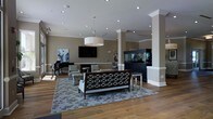 This refined single-floor space presents an elegant and welcoming environment designed for comfort and sophistication. The open-plan layout is anchored by rich hardwood flooring and high ceilings adorned with recessed lighting and a statement drum pendant chandelier, creating a bright and airy atmosphere throughout.

Structural white columns with classic crown molding define distinct areas within the space while maintaining an open, flowing feel. A large flat-screen television is mounted prominently on a taupe accent wall, flanked by tasteful artwork, including a vibrant blue abstract painting that adds a pop of color to the neutral palette.

The seating area features an elegant lattice-back bench settee positioned atop a patterned area rug, complemented by a pair of upholstered accent chairs arranged around a side table — ideal for relaxed conversation or leisure. A grand piano positioned near natural light from a side window adds a cultural and artistic touch to the ambiance.

A striking large aquarium encased in a dark cabinetry surround serves as a dramatic focal point, adding a serene, living element to the space. The overall design balances warmth and refinement, with a neutral taupe and cream color palette, quality furnishings, and thoughtful architectural details that together create an inviting and polished environment.