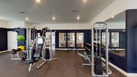 Located at 7220 E Broad St in Columbus, Ohio, this fitness center offers a dedicated, single-floor space designed to support a comprehensive workout routine. The gym is outfitted with a solid selection of strength training equipment, including cable weight machines, adjustable benches, and a full dumbbell rack, providing users with a wide range of exercise options for both targeted and full-body training.

The interior features a bold and energizing design, with deep navy blue accent walls that contrast sharply against the light-colored upper walls and smooth white ceiling. Recessed lighting illuminates the space evenly, ensuring good visibility throughout the workout area. The flooring consists of durable rubber-style gym matting, well-suited for heavy equipment and high-activity use.

A wall of mirrors runs along one side of the room, visually expanding the space while allowing users to monitor their form during exercise. The mirrors also frame a view into an adjacent room, which appears to be a lounge or common area, adding a sense of openness and connectivity to the overall layout. The space is clean, organized, and purposefully arranged to maximize functionality, making it a practical and well-appointed facility for residents or users of the property.