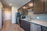 Located at 201 E Main St in the heart of Waunakee, Wisconsin, this efficiently designed one-bedroom, one-bathroom unit offers a thoughtfully arranged single-floor layout with modern finishes throughout. The open-concept kitchen and living area serves as the central hub of the space, featuring rich dark-stained cabinetry, white quartz-style countertops, a full suite of stainless steel appliances including a refrigerator, range, microwave, and dishwasher, and a subway tile backsplash. Warm hardwood-style luxury vinyl plank flooring runs throughout the main living areas, complemented by neutral gray walls that lend a clean, contemporary feel. A hallway with a sliding barn door connects the main living area to the private bedroom, which is carpeted for comfort and includes access to a dedicated closet. The full bathroom features a dark wood vanity with an undermount sink, white countertop, and a well-lit mirror. An in-unit laundry area adds everyday convenience. A private balcony with cable railing offers open views of downtown Waunakee's streetscape and surrounding buildings, bringing in natural light and a connection to the vibrant community below.