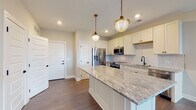 Located at 1550 Kenny Rd in Columbus, Ohio, this single-floor residence offers a thoughtfully designed open-concept layout with high-quality finishes throughout. The kitchen serves as the heart of the home, showcasing crisp white shaker cabinetry, a striking granite-topped center island illuminated by a pair of brass pendant lights, and a herringbone-patterned marble tile backsplash. Stainless steel appliances — including a French door refrigerator, range, over-the-range microwave, and dishwasher — provide a full suite of modern amenities. A matte black faucet at the undermount sink adds a refined contrast to the bright cabinetry. Rich hardwood-style flooring flows throughout the main living areas, connecting the kitchen seamlessly to the living space, which benefits from natural light through large windows and access to an outdoor balcony. The bathroom is appointed with an eye-catching geometric hexagon tile floor, deep navy subway tile accents, a granite-topped vanity with matte black fixtures, and gold-toned sconce lighting. A spacious walk-in closet with wire shelving systems and carpeted flooring provides generous storage. The overall palette of soft neutrals, warm metallics, and bold tile accents gives the space a contemporary yet welcoming character.