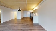 This single-floor unit showcases a thoughtfully designed open layout that seamlessly connects the living and kitchen areas. A striking exposed wood-panel ceiling runs throughout, lending a warm, contemporary character to the space and creating a cohesive aesthetic across rooms. The living area features smooth hardwood-style flooring, large windows dressed with sheer curtains, and a wall-mounted HVAC unit, while a sliding door provides access to an outdoor balcony with views of the surrounding neighborhood and distant landscape.

The kitchen is well-appointed with stainless steel appliances, including a refrigerator, electric range, and dishwasher. Taupe-toned cabinetry is complemented by open metal shelving, a tile backsplash, and clean light countertops, delivering a functional and modern workspace. A bold pendant light anchors the open-plan kitchen and living zone.

The bedrooms are carpeted for comfort and feature wall sconce lighting and windows that bring in natural light. One bedroom offers a scenic outdoor view, while another provides a more private setting. A bathroom is also present within the unit. Overall, the space balances warmth and minimalism through its consistent use of natural wood tones, clean white walls, and modern finishes throughout.
