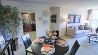 This single-floor residence showcases an open-concept design that seamlessly integrates the dining area, living room, and kitchen. The dining space is anchored by a round dark-finished table paired with matching dark chairs, set atop neutral-toned carpeting that extends throughout the main living areas. Decorative wall accents, including circular botanical-style pieces, add visual interest to the soft, neutral walls.

The adjacent living area provides a comfortable extension of the space, furnished with a light-toned sofa and floor lamp, creating a relaxed yet refined atmosphere. A framed artwork piece on the far wall adds a finishing touch to the living zone.

The kitchen, visible through an open passageway, features rich dark-toned cabinetry with ample storage, a dark refrigerator, and a functional sink with a window above it, allowing natural light to filter in. The warm cabinetry tones contrast effectively with the neutral surroundings.

Overall, the layout promotes easy flow between areas, making it practical for both everyday living and entertaining. The consistent neutral color palette and coordinated furnishings contribute to a cohesive, inviting ambiance throughout.