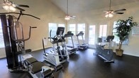This fitness center is designed to provide a functional and comfortable workout experience within a single-level layout. The space features soaring vaulted ceilings that create an open, airy atmosphere, complemented by two ceiling fans that ensure steady airflow throughout the room. Large windows line the walls, flooding the interior with generous natural light and offering views to the outdoors, while a set of glass-paneled doors provides direct access to an exterior area, further enhancing the connection to the outside environment. The flooring is finished in durable dark rubber matting, ideal for high-traffic exercise use and equipment placement. The facility is outfitted with a well-rounded selection of cardio equipment, including multiple treadmills, elliptical cross-trainers, and a stationary recumbent bike, all from quality fitness brands. Additional strength and stretching equipment is also present. The bright, clean aesthetic of the neutral walls combined with the expansive windows creates an energizing and welcoming environment suited for a variety of fitness routines. Overall, the space strikes a balance between practicality and comfort, making it an inviting destination for daily exercise.
