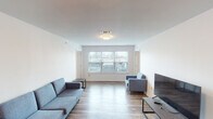 This single-floor unit offers a clean, functional layout designed for comfortable everyday living. The living area is bright and open, featuring wood-look flooring, white walls, and a large multi-pane window that fills the space with natural light and provides views of the surrounding exterior. The room comfortably accommodates a sofa, accent chair, and television setup, giving it a practical, welcoming feel.

The kitchen is equipped with warm-toned wood cabinetry, light countertops, and a full suite of stainless steel appliances including a range, dishwasher, microwave, and refrigerator. A range hood is positioned above the stove, and the layout allows for efficient use of the cooking space. A hallway connects the kitchen to the rest of the unit, leading to the main entrance door.

The bedroom features carpeted flooring, white walls, and windows on two sides that bring in ample natural light. An adjacent closet provides additional storage. The full bathroom includes wood-look flooring, a soaking tub, toilet, vanity with a wood cabinet, and a grab bar, indicating accessibility-conscious design. A dedicated laundry closet houses a full-size front-loading washer and dryer set, completing the unit's practical amenities.