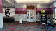 This single-floor fitness center is thoughtfully designed to support a complete workout experience. The space is immediately recognizable by its distinctive tri-tone wall design, featuring alternating bands of burgundy, gray, and charcoal that lend the facility a bold, energetic atmosphere throughout.

The main cardio area is a standout feature, lined with multiple treadmills and elliptical machines, each equipped with individual screens for entertainment during workouts. Large windows along the cardio wall flood the space with natural light and offer views of the surrounding outdoor landscape. Full-length mirrors on adjacent walls visually expand the room and assist with form checks during exercise.

A dedicated spin and functional training area houses stationary cycling bikes alongside a well-stocked rack of medicine balls, stability balls, and resistance equipment, catering to a variety of training styles. Wall hooks near the entrance provide convenient storage for jump ropes and resistance bands.

The facility also includes a separate package locker room, lined floor-to-ceiling with secure parcel lockers and featuring its own exterior access door—a practical amenity for residents. Patterned carpet tiles in muted red and gray tones anchor each zone of the facility, tying the design together cohesively. Recessed lighting throughout ensures consistent illumination across all areas of the space.