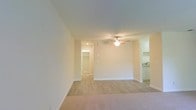 This single-floor space presents a clean, functional layout centered around an open living area finished in warm neutral tones. The room is anchored by plush wall-to-wall carpeting that adds comfort underfoot, while smooth, light-colored walls and a standard ceiling height contribute to a bright and airy atmosphere. A ceiling fan with integrated lighting serves as the focal point overhead, providing both air circulation and ambient illumination throughout the space.

Along the far wall, a doorway leads to what appears to be a dedicated closet or storage area, complete with built-in shelving and a mirrored door, offering practical organizational solutions. To the right side of the living area, a partial view into the kitchen reveals white cabinetry and a sink, suggesting a well-appointed culinary space with classic finishes. The transition between the carpeted living area and what appears to be hard flooring in the kitchen is clearly defined, adding visual distinction between the two zones.

Overall, the space maintains a simple, well-maintained aesthetic with consistent neutral tones throughout, making it a versatile and move-in ready environment.