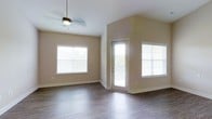 This single-floor living space offers a clean, open layout defined by its vaulted ceilings that lend a sense of spaciousness and airiness to the interior. The main living area is finished with rich, dark hardwood-style flooring that contrasts attractively with the warm beige-toned walls, creating a welcoming and cohesive aesthetic throughout.

Natural light is a standout feature of this space, with multiple large windows fitted with horizontal blinds allowing for adjustable brightness. A glass-paneled entry door further enhances the flow of natural light into the interior while providing a visual connection to the outdoors. The windows are positioned to illuminate the room from multiple angles, reducing the need for artificial lighting during daylight hours.

A ceiling fan with an integrated light fixture is centrally mounted, offering both functional air circulation and supplemental lighting. The clean, neutral color palette of the walls and trim provides a versatile backdrop suitable for a variety of interior design styles. The combination of vaulted ceilings, generous natural light, and open floor plan gives this space a bright, airy, and move-in-ready feel.