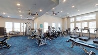 This fitness center is designed to support a well-rounded exercise routine, offering a broad selection of both strength training and cardiovascular equipment. The space is outfitted with cable weight machines, seated resistance equipment, ellipticals, stationary bikes, and treadmills, providing options for users of all fitness levels. The room features vaulted ceilings that lend a sense of openness, while multiple ceiling fans are evenly distributed throughout, ensuring consistent airflow and comfort during workouts. Recessed lighting supplements the natural light that enters through several windows positioned along the walls, keeping the interior bright and welcoming. The flooring consists of durable blue-toned carpet tiles, which offer cushioning underfoot while adding a modern visual element to the space. The neutral wall tones and clean layout contribute to an uncluttered, functional atmosphere. Overall, the fitness center is thoughtfully arranged to maximize usable floor space while maintaining clear circulation paths between equipment stations, making it a practical and accessible amenity for regular use.