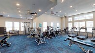 This fitness center is designed to support a well-rounded exercise routine, offering a broad selection of both strength training and cardiovascular equipment. The space is outfitted with cable weight machines, seated resistance equipment, ellipticals, stationary bikes, and treadmills, providing options for users of all fitness levels. The room features vaulted ceilings that lend a sense of openness, while multiple ceiling fans are evenly distributed throughout, ensuring consistent airflow and comfort during workouts. Recessed lighting supplements the natural light that enters through several windows positioned along the walls, keeping the interior bright and welcoming. The flooring consists of durable blue-toned carpet tiles, which offer cushioning underfoot while adding a modern visual element to the space. The neutral wall tones and clean layout contribute to an uncluttered, functional atmosphere. Overall, the fitness center is thoughtfully arranged to maximize usable floor space while maintaining clear circulation paths between equipment stations, making it a practical and accessible amenity for regular use.