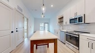 Located at 2100 Marquis Pkwy in Williamsburg, Virginia, this one-bedroom, one-bathroom unit offers a thoughtfully designed layout across a single floor. The open-concept kitchen and living area forms the heart of the space, finished with light wood-style flooring that extends throughout the main areas. The kitchen features white shaker-style cabinetry with matte black hardware, a subway tile backsplash, open wood shelving, stainless steel appliances including a range, microwave, dishwasher, and refrigerator, and a large island with a white countertop that doubles as a dining and prep area. Pendant lighting with glass fixtures adds a modern accent overhead. The living area benefits from natural light through windows and a glazed exterior door. The bedroom is generously sized with plush carpeting, a ceiling fan, and a window with exterior views. A dedicated walk-in closet provides ample storage with wire shelving systems. The bathroom is clean and contemporary, featuring a white vanity with a quartz-style countertop, a large framed mirror, and a tub/shower combo with subway tile surround and brushed nickel fixtures.