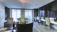 This single-floor space combines a stylish lounge area with a functional business center, creating a versatile environment suited for both relaxation and productivity. The lounge section is furnished with plush, earth-toned armchairs and sofas accented by patterned throw pillows, arranged to encourage comfortable conversation or quiet lounging. Large floor-to-ceiling windows flood the seating area with abundant natural light while offering views of an outdoor courtyard, seamlessly connecting the interior to the exterior environment. The flooring transitions between dark hardwood in the lounge zone and large-format gray tiles, subtly delineating the different functional areas. A dedicated business center runs along one wall, featuring a sleek, built-in desk with computer workstations and modern office chairs, framed by dark, textured wall panels that add a sophisticated contrast to the lighter tones throughout the rest of the space. Track lighting overhead supplements the natural light, ensuring the entire area is well-illuminated. The overall ambiance is polished and contemporary, balancing professional utility with a welcoming, residential comfort.