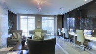 This single-floor space combines a stylish lounge area with a functional business center, creating a versatile environment suited for both relaxation and productivity. The lounge section is furnished with plush, earth-toned armchairs and sofas accented by patterned throw pillows, arranged to encourage comfortable conversation or quiet lounging. Large floor-to-ceiling windows flood the seating area with abundant natural light while offering views of an outdoor courtyard, seamlessly connecting the interior to the exterior environment. The flooring transitions between dark hardwood in the lounge zone and large-format gray tiles, subtly delineating the different functional areas. A dedicated business center runs along one wall, featuring a sleek, built-in desk with computer workstations and modern office chairs, framed by dark, textured wall panels that add a sophisticated contrast to the lighter tones throughout the rest of the space. Track lighting overhead supplements the natural light, ensuring the entire area is well-illuminated. The overall ambiance is polished and contemporary, balancing professional utility with a welcoming, residential comfort.