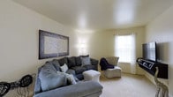 This two-floor residence offers a thoughtfully designed layout that balances functionality with a warm, inviting ambiance. The living room serves as a central gathering space, highlighted by vaulted ceilings that lend a sense of openness and volume to the area. Plush wall-to-wall carpeting in a neutral tone runs underfoot, contributing to the room's cozy atmosphere. A large L-shaped sectional sofa in a deep charcoal gray anchors the space, complemented by an accent armchair with a subtle botanical print and a stylish upholstered ottoman. Soft, warm lighting from a tall floor lamp enhances the relaxed feel of the room. Natural light filters in through a window dressed with white curtains and blinds, adding brightness to the neutral-toned walls. Decorative framed artwork featuring nature-inspired motifs adorns the walls, adding visual interest and a cohesive aesthetic throughout the living area. The overall design of the space reflects a calm, understated style with carefully chosen furnishings that prioritize comfort. The two-floor configuration provides ample room for a variety of living arrangements, making this a versatile and well-appointed home.