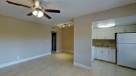 This single-floor space presents an open and functional layout, finished in neutral warm-toned walls that create a welcoming, understated atmosphere throughout. The main living area is finished with light-toned ceramic tile flooring that extends seamlessly across the space, contributing to a bright and cohesive feel. Overhead, a ceiling fan with an integrated light fixture provides both air circulation and ambient lighting, while a track lighting system mounted along the ceiling adds directed illumination to the living area.

A partial dividing wall separates the living space from the kitchen, which is positioned to the side and features white cabinetry both above and below the counter. The kitchen includes a sink, a white range, and a tile backsplash that adds a practical, clean detail. An overhead flush-mount light fixture brightens the kitchen area effectively.

A hallway visible from the main living space leads toward additional rooms, suggesting a practical floor plan with private areas set apart from the main living zone. White baseboards frame the rooms cleanly, complementing the neutral palette. Overall, the space offers a straightforward, functional interior with a calm, livable ambiance.