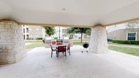 This outdoor communal space offers a well-appointed covered patio designed for relaxed gathering and al fresco dining. The area is anchored by a robust stone column that supports a solid overhead canopy, providing ample shade and shelter for residents and guests. A dining table with chairs sits centrally beneath the cover, creating an inviting setting for shared meals and socializing.

Grilling amenities are a standout feature of this space, with built-in gas grills housed in a dedicated outdoor kitchen station, complemented by a freestanding charcoal kettle grill nearby — offering flexibility for various cooking preferences. The concrete flooring provides a clean, durable surface throughout the covered section.

Beyond the shaded patio, the surrounding grounds open up to well-maintained green lawns and mature trees that add natural character to the environment. The broader residential community is visible in the background, with multi-story buildings and additional landscaping contributing to the overall neighborhood feel. The open sightlines and greenery create a pleasant, park-like ambiance that enhances the usability of this outdoor amenity space throughout the day.