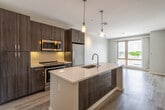 This contemporary single-floor unit showcases a thoughtfully designed open-concept layout that seamlessly connects the kitchen and living areas. The kitchen serves as the centerpiece, anchored by a large island with striking quartz countertops featuring subtle speckled detailing and an undermount stainless steel sink with a pull-down faucet. Pendant lighting hangs above the island, providing focused illumination and a touch of modern elegance. Dark wood-toned cabinetry extends floor-to-ceiling, offering generous storage and a bold visual contrast against the light countertops. A full suite of stainless steel appliances, including a refrigerator, range, and over-the-range microwave, completes the kitchen.

The living area benefits from abundant natural light through large, floor-to-ceiling windows that offer views of the surrounding neighborhood. Light wood-style flooring runs throughout the main living spaces, contributing to a warm, cohesive aesthetic. Neutral gray walls maintain a clean, versatile backdrop throughout the unit.

The bedroom features a tall, narrow window that frames an outdoor view, a double louvered closet for storage, and the same wood-style flooring found in the main areas. The bathroom is finished with large-format tile flooring, a tub-shower combination with tiled surround, a dark wood vanity, and clean, minimalist fixtures, rounding out this well-appointed space.