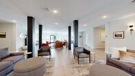 Located on Legends Drive in Champaign, Illinois, this single-floor community lounge space offers a thoughtfully designed, open-plan layout that encourages gathering and relaxation. The interior is characterized by light hardwood-style flooring, recessed lighting, and a neutral color palette of whites and grays, accented by bold dark structural columns that add a striking architectural contrast throughout the space.

Upon entry, a welcoming foyer greets visitors with large glass doors and floor-to-ceiling windows that flood the space with natural light and offer views of the surrounding area. The main lounge area is expansive and divided into multiple distinct seating zones, each furnished with a mix of contemporary sofas, leather armchairs, and accent tables arranged on layered area rugs. A standout focal point is the tiled fireplace, topped by a mounted television and flanked by comfortable seating, creating a warm and inviting gathering area.

Decorative wall art, potted plants, and carefully curated accessories contribute to the space's modern yet approachable aesthetic. Additional rooms accessible from the main lounge provide further utility and privacy. Throughout, the design balances openness with defined zones, making this a versatile and well-appointed communal space suited for socializing and leisure.
