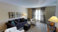 This two-floor residence offers a thoughtfully arranged living space with a warm, neutral color palette that creates a comfortable and inviting atmosphere. The main living area is carpeted and generously proportioned, providing ample room for varied furniture arrangements. A large window dressed with bold, black-and-white botanical-patterned curtains serves as a focal point, allowing natural light to brighten the space throughout the day.

The living room is furnished with a deep navy sofa accented by teal and dark throw pillows, complemented by a white accent chair with a patterned cushion and a black leather storage ottoman. A floor lamp adds warm ambient lighting, while two decorative wall art panels displaying intricate botanical motifs add visual interest to the neutral beige walls. A small potted plant near the window introduces a touch of greenery to the room.

The overall aesthetic balances casual comfort with coordinated décor, making the living area a practical yet stylish environment. The two-floor layout suggests additional living, sleeping, or functional spaces above, offering versatility and separation between different areas of the home.