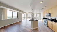 This single-floor residence is situated at an elevated position within a high-rise building, offering expansive views of the surrounding cityscape and greenery through generous floor-to-ceiling windows. The open-concept layout seamlessly connects the living and kitchen areas, creating a bright, airy atmosphere filled with abundant natural light throughout the day.

The kitchen is thoughtfully designed with light maple cabinetry extending to the ceiling, dark granite countertops, a stainless steel appliance suite including a gas range, built-in microwave, and refrigerator, and a central island with an undermount sink and pendant lighting above. The transition from the kitchen's tile flooring to the rich hardwood of the living area clearly delineates the two spaces while maintaining a cohesive aesthetic.

The living area is expansive and flooded with light, featuring gleaming hardwood floors and multiple large windows framing elevated outdoor views. The bedroom offers plush carpeting, a sliding glass door leading to a private balcony, and a well-sized closet with wire shelving. The bathrooms are finished with light maple vanities, white stone countertops, large-format tile flooring, and a full soaking tub with dark tile surround.