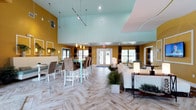 This single-floor community clubhouse is designed with a bold, contemporary aesthetic, highlighted by warm golden-yellow walls, soft teal ceilings, and herringbone-patterned wood-look tile flooring throughout the main areas. The expansive open-plan lounge and dining space is the centerpiece of the facility, boasting soaring vaulted ceilings, dramatic track lighting, and pendant fixtures that draw the eye upward. A large white sectional sofa and a wall-mounted flat-screen TV anchor the lounge area, while a long communal dining table with upholstered chairs provides generous seating for gatherings. Abundant natural light filters in through multiple windows and glass French doors dressed with striped curtain panels. The kitchen is well-appointed with white cabinetry, dark countertops, a teal tile backsplash, stainless steel appliances including a full-size refrigerator, and a breakfast bar with seating. Adjacent to the main area, the business center offers a row of desktop computers along a built-in workstation, complemented by a sleek glass conference table surrounded by modern white ergonomic chairs, recessed lighting, and colorful artwork on the walls. A separate conference room provides a more private meeting environment with its own table, chairs, and natural light from large windows. The overall ambiance is energetic yet functional, blending vivid color with modern furnishings.