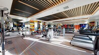 This fitness center is a thoughtfully designed, single-level facility that combines vibrant aesthetics with comprehensive workout amenities. The space is divided into clearly defined zones, each tailored to specific training needs. A striking design feature throughout is the bold, multicolored geometric accent wall in red, yellow, navy, and white, which energizes the environment and serves as a visual anchor across multiple areas of the gym.

The ceiling is a standout architectural element, featuring a layered slatted wood and metal batten design that adds warmth and visual depth while contributing to the overall modern character of the space. Recessed lighting complements abundant natural light that streams through large floor-to-ceiling windows, offering views of an outdoor pool and patio area with palm trees.

The strength training zone is generously equipped with cable machines, weight racks, barbells, and benches arranged on hardwood-style flooring. A separate cardio area houses rows of treadmills, rowing machines, and upright bikes, positioned to take advantage of the outdoor views. A glass-partitioned studio space accommodates cycling equipment against vibrant red-accented walls. Full-length mirrors amplify the sense of space throughout, and striped carpet flooring in the cardio zone adds a subtle design detail that ties into the facility's energetic color palette.
