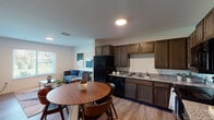 This single-floor residence is thoughtfully designed with a cohesive, modern aesthetic carried throughout its open layout. The kitchen serves as a functional centerpiece, outfitted with rich dark-toned cabinetry, granite countertops, and black appliances including a refrigerator, dishwasher, range, and over-the-range microwave. Light wood-style flooring runs throughout the kitchen and adjoining dining area, where space for a round dining table sits comfortably near the entry. The open-concept design allows the kitchen to flow naturally into the living room, which receives generous natural light through large windows fitted with horizontal blinds offering outdoor views of surrounding greenery. The living area provides a welcoming, relaxed atmosphere well-suited for everyday use. The bedroom continues the natural light theme with a sizable window, ceiling fan, and a roomy closet with built-in shelving. The bathroom features marble-patterned surround walls on the tub and shower, tile flooring, and accessibility-conscious grab bars throughout. Overall, the residence presents a clean, practical, and inviting living environment.