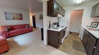 This single-story, one-bedroom, one-bathroom residence offers a thoughtfully arranged layout that maximizes functionality and comfort. The galley-style kitchen is a standout feature, outfitted with dark espresso cabinetry lining both walls, white countertops, a white electric range, refrigerator, and a built-in sink with ample storage above and below. Wood-look flooring runs through the kitchen, adding warmth and contrast to the cabinetry. The kitchen opens toward the living area, creating a connected flow between spaces.

The living area is carpeted and benefits from large sliding windows that frame lush, green outdoor views, filling the room with natural light and creating a welcoming ambiance. The bedroom continues this bright, airy feel with its own generous window overlooking the outdoors, plush carpet flooring, and neutral wall tones that lend a calm, restful atmosphere.

The bathroom features a wide vanity with a light-toned countertop, a large mirror, white cabinetry, a tub-shower combination, and a window that allows for natural ventilation. Throughout the unit, the overall design emphasizes clean lines, neutral tones, and practical living spaces suited for everyday comfort.