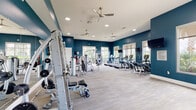This single-floor fitness center offers a comprehensive workout environment designed to accommodate a variety of exercise routines. The space is defined by bold teal accent walls that contrast with crisp white ceilings, creating an energetic and motivating atmosphere. Light wood-style flooring runs throughout, lending a clean, modern aesthetic to the interior.

The main workout area is generously proportioned, housing an extensive selection of equipment that includes multiple treadmills, elliptical trainers, stationary bikes, and a multi-station cable and weight machine setup. Free weights, resistance equipment, and stability balls are also available, catering to both strength and functional training needs. Wall-mounted televisions are positioned throughout the space for entertainment during workouts.

Large windows line the walls, flooding the interior with natural light while offering views of the surrounding outdoor landscape, including palm trees and open greenery. Ceiling fans are mounted overhead to maintain air circulation throughout the space. A fitness vending machine provides convenient access to snacks and supplements. An accessible restroom with tile flooring and grab bars is located adjacent to the main fitness area.