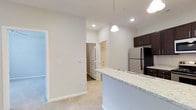 This single-floor residence offers a clean, modern aesthetic throughout its open-concept layout. The kitchen is a standout feature, equipped with rich dark cabinetry, stainless steel appliances including a full-size refrigerator, and a generous peninsula counter finished with light granite-style stone. A pendant light hangs above the counter, adding a refined touch to the space. The kitchen flows seamlessly into the adjacent living area, which is finished with plush carpeting and neutral white walls that contribute to a bright, airy atmosphere. Recessed lighting throughout keeps the space well-illuminated. A hallway leads toward a bathroom, partially visible in the background, which features a bathtub and shower combination. The doors throughout are classic paneled white, maintaining a consistent and cohesive interior style. The overall ambiance is calm and neutral, with a move-in-ready feel that allows for a variety of furnishing styles. High ceilings add a sense of openness and volume to the space, enhancing the comfortable and functional living environment presented across the single floor.