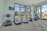 Located at 36125 Creighton Avenue in Murrieta, CA, this single-floor fitness center offers a generously sized, thoughtfully designed workout facility divided into two distinct exercise rooms. The main exercise room is defined by expansive floor-to-ceiling windows with sleek black frames that flood the space with natural light and provide open views of the surrounding community and hills. This room features a checkered tile floor and is outfitted with a comprehensive selection of cardio and strength training equipment, including stationary bikes, cable machines, rowing equipment, and weight benches. Ceiling fans circulate air throughout, enhancing comfort during workouts. A secondary exercise room, finished with warm yellow accent walls and light wood-style flooring, offers a more versatile training environment. This space includes free weights, medicine balls, resistance equipment, and additional cardio machines. Large windows continue to bring in natural light, and wall-mounted mirrors enhance the sense of openness. Glass double doors provide direct access to the outdoor area. Together, the two rooms create a well-rounded fitness facility that caters to a variety of workout preferences in a bright, modern, and welcoming environment.