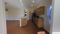 This single-floor home showcases a thoughtfully designed open-concept layout that seamlessly connects the kitchen and living areas, creating a spacious and welcoming environment. The kitchen is a standout feature, equipped with stainless steel appliances — including a refrigerator, range, and over-the-range microwave — complemented by warm honey-toned wooden cabinetry and granite countertops. A central kitchen island with matching granite and decorative column accents provides additional prep space and serves as a natural gathering point. Rich, dark hardwood flooring flows throughout, adding warmth and visual continuity across the space. Vaulted ceilings rise above the open living and kitchen area, enhancing the sense of volume and airiness. Recessed lighting is strategically placed throughout the ceiling, ensuring consistent and functional illumination. Neutral, light-toned walls contribute to a clean and versatile aesthetic. A doorway visible from the main area suggests additional rooms beyond, offering a sense of depth and privacy. The overall ambiance is practical yet polished, making this home well-suited for everyday living and casual entertaining alike.