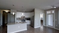 This single-floor residence on 9th Street in Durham, North Carolina, showcases a clean, modern aesthetic with an open-concept layout that promotes a sense of spaciousness and flow. The kitchen serves as a central highlight, featuring white shaker-style cabinetry that extends to the ceiling, complemented by a gray subway tile backsplash and light granite countertops. A peninsula island provides additional prep space and casual seating, while pendant lighting adds a stylish focal point overhead. Stainless steel appliances, including a gas range, microwave, and full-size refrigerator, complete the well-equipped cooking space. The flooring throughout is a warm-toned hardwood laminate that ties the living areas together cohesively. Neutral gray walls maintain a versatile, contemporary palette throughout. A doorway visible from the kitchen leads to an additional room that benefits from natural light through a window, suggesting a well-connected floor plan. The overall ambiance is modern and move-in ready, with consistent finishes and thoughtful design details that reflect current residential design standards.