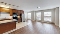 This single-floor residence offers a thoughtfully designed open-concept layout that seamlessly connects the kitchen and living area. The kitchen is a standout feature, equipped with rich medium-toned wood cabinetry extending to the ceiling for ample storage, granite countertops with subtle earth-tone veining, and a full suite of stainless steel appliances including a refrigerator, electric range, over-the-range microwave, and dishwasher. Light tile flooring anchors the kitchen zone, transitioning into warm hardwood-style flooring throughout the expansive living area. Large windows along the living space admit generous natural light and provide views of the surrounding urban streetscape. Track lighting and flush-mount ceiling fixtures provide additional illumination throughout. The primary bathroom features a granite-topped vanity, tile-surround soaking tub, and light tile flooring, while a second bathroom offers a similar granite vanity and a tiled shower. The bedrooms are carpeted for comfort, each featuring neutral wall tones, natural light from windows, and closet space with built-in shelving. Overall, the unit presents a clean, modern aesthetic with consistent finishes throughout.