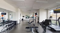 This modern fitness center is thoughtfully designed to support a comprehensive workout experience across a single open floor. The space is bright and clean, featuring neutral white walls, a drop-panel ceiling with recessed lighting, and durable grey flooring throughout.

The cardio zone is positioned along a wall of large floor-to-ceiling windows that offer expansive views of the surrounding landscape, creating an energizing atmosphere for treadmill, elliptical, and stationary bike workouts. Multiple flat-screen TVs are mounted throughout the space for entertainment during exercise.

A dedicated free weights area houses a full rack of dumbbells in varying sizes, while a multi-station cable and strength machine provides options for a wide range of resistance training exercises. Adjustable benches are available throughout the floor for versatile use.

The functional training zone features exercise balls, kettlebells, resistance bands, and yoga mats, supported by a large wall-mounted mirror that spans a significant portion of the room — ideal for form monitoring during workouts. Modular storage cubbies keep accessories organized and accessible.

Practical amenities include a hand sanitizer station, waste receptacles, and a water station near the entrance. The overall layout is open and spacious, allowing for easy movement between equipment zones while maintaining a clean, professional aesthetic.
