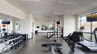 This modern fitness center is thoughtfully designed to support a comprehensive workout experience across a single open floor. The space is bright and clean, featuring neutral white walls, a drop-panel ceiling with recessed lighting, and durable grey flooring throughout.

The cardio zone is positioned along a wall of large floor-to-ceiling windows that offer expansive views of the surrounding landscape, creating an energizing atmosphere for treadmill, elliptical, and stationary bike workouts. Multiple flat-screen TVs are mounted throughout the space for entertainment during exercise.

A dedicated free weights area houses a full rack of dumbbells in varying sizes, while a multi-station cable and strength machine provides options for a wide range of resistance training exercises. Adjustable benches are available throughout the floor for versatile use.

The functional training zone features exercise balls, kettlebells, resistance bands, and yoga mats, supported by a large wall-mounted mirror that spans a significant portion of the room — ideal for form monitoring during workouts. Modular storage cubbies keep accessories organized and accessible.

Practical amenities include a hand sanitizer station, waste receptacles, and a water station near the entrance. The overall layout is open and spacious, allowing for easy movement between equipment zones while maintaining a clean, professional aesthetic.