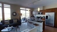 This contemporary single-floor space showcases a thoughtfully designed open-concept layout that seamlessly integrates the kitchen, dining, and living areas into one cohesive environment. The kitchen is a standout feature, equipped with stainless steel appliances including a range and built-in microwave, rich warm-toned cabinetry, and a striking dark granite or stone breakfast bar with modern bar stools. Pendant lighting hangs above the kitchen island, adding both function and style. The living area is generously proportioned, offering a comfortable, modern aesthetic with neutral tones, a plush sofa, and accent armchairs. A decorative sunburst mirror and floor lamp contribute to the curated, contemporary feel of the space. Large floor-to-ceiling sliding glass doors and expansive windows flood the interior with abundant natural light while providing direct access to an outdoor balcony, where outdoor seating captures elevated open views of the surrounding cityscape. Track lighting on the ceiling complements the natural light throughout the day. The overall ambiance is polished and urban, blending practical design with modern aesthetics to create a welcoming and functional living environment.