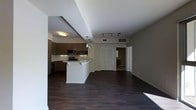 This single-floor space showcases a thoughtfully designed open-concept layout that seamlessly connects the kitchen and living areas. The kitchen is outfitted with dark espresso cabinetry, stainless steel appliances including a microwave and range, and a light-colored countertop island with an undermount sink — a functional centerpiece ideal for both meal preparation and casual dining. Pendant lighting hangs above the island, complementing the flush-mount ceiling fixture and adding warmth to the cooking space. A tile backsplash adds a refined touch to the kitchen's design.

The living area extends generously from the kitchen, featuring rich dark hardwood flooring that runs throughout the entire space, providing a cohesive and elegant foundation. Track lighting along the ceiling offers flexible illumination across the open floor plan. White walls and bright trim keep the interior feeling clean and airy.

Double doors lead to what appears to be a closet or additional storage, while a hallway beyond suggests access to a bedroom and bathroom. The overall ambiance is contemporary and polished, making for a sleek and functional living environment.