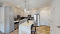 This thoughtfully designed single-floor unit at 3100-34 S 7th St in St. Louis, MO, combines contemporary finishes with warm, textural accents throughout. The open-concept kitchen is a standout feature, showcasing gray shaker cabinetry, a bold herringbone tile backsplash, stainless steel appliances, and a large waterfall-edge quartz island with bar seating. Geometric pendant lights add a modern touch overhead, while light hardwood flooring flows seamlessly into the adjoining living area.

The living room is anchored by a dramatic stacked-stone accent wall, creating a striking contrast against the soft gray painted walls and natural light streaming through large windows. A geometric chandelier echoes the kitchen's pendant fixtures, maintaining visual cohesion throughout the open layout.

The bedrooms feature plush carpeting and large windows with roller shades and curtains, one bedroom highlighting its own stacked-stone feature wall. Two bathrooms are finished with dark large-format tile flooring, white subway tile, gray shaker vanities with white countertops, and clean brushed-nickel fixtures—one with a walk-in shower and one with a tub-shower combination.