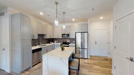 This thoughtfully designed single-floor unit at 3100-34 S 7th St in St. Louis, MO, combines contemporary finishes with warm, textural accents throughout. The open-concept kitchen is a standout feature, showcasing gray shaker cabinetry, a bold herringbone tile backsplash, stainless steel appliances, and a large waterfall-edge quartz island with bar seating. Geometric pendant lights add a modern touch overhead, while light hardwood flooring flows seamlessly into the adjoining living area.

The living room is anchored by a dramatic stacked-stone accent wall, creating a striking contrast against the soft gray painted walls and natural light streaming through large windows. A geometric chandelier echoes the kitchen's pendant fixtures, maintaining visual cohesion throughout the open layout.

The bedrooms feature plush carpeting and large windows with roller shades and curtains, one bedroom highlighting its own stacked-stone feature wall. Two bathrooms are finished with dark large-format tile flooring, white subway tile, gray shaker vanities with white countertops, and clean brushed-nickel fixtures—one with a walk-in shower and one with a tub-shower combination.